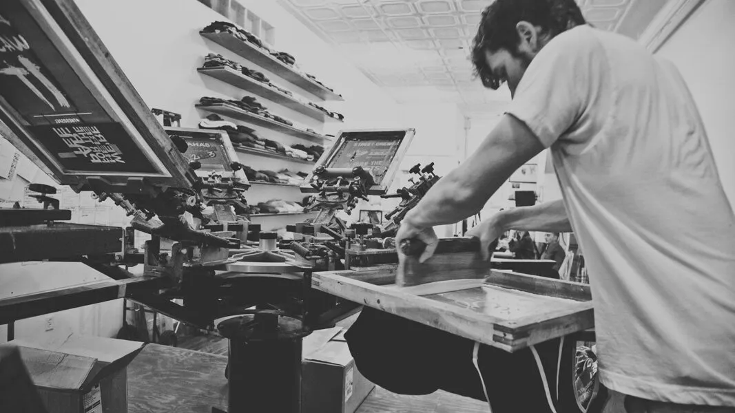 Black-and-white photo of a printer pulling a squeegee across a screen on a manual screen printing press