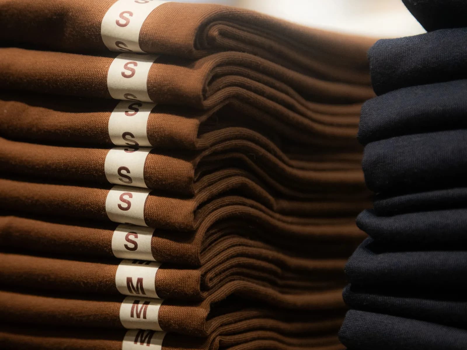 Stacks of identical folded brown shirts with size strips marked S and M, beside a stack of black shirts