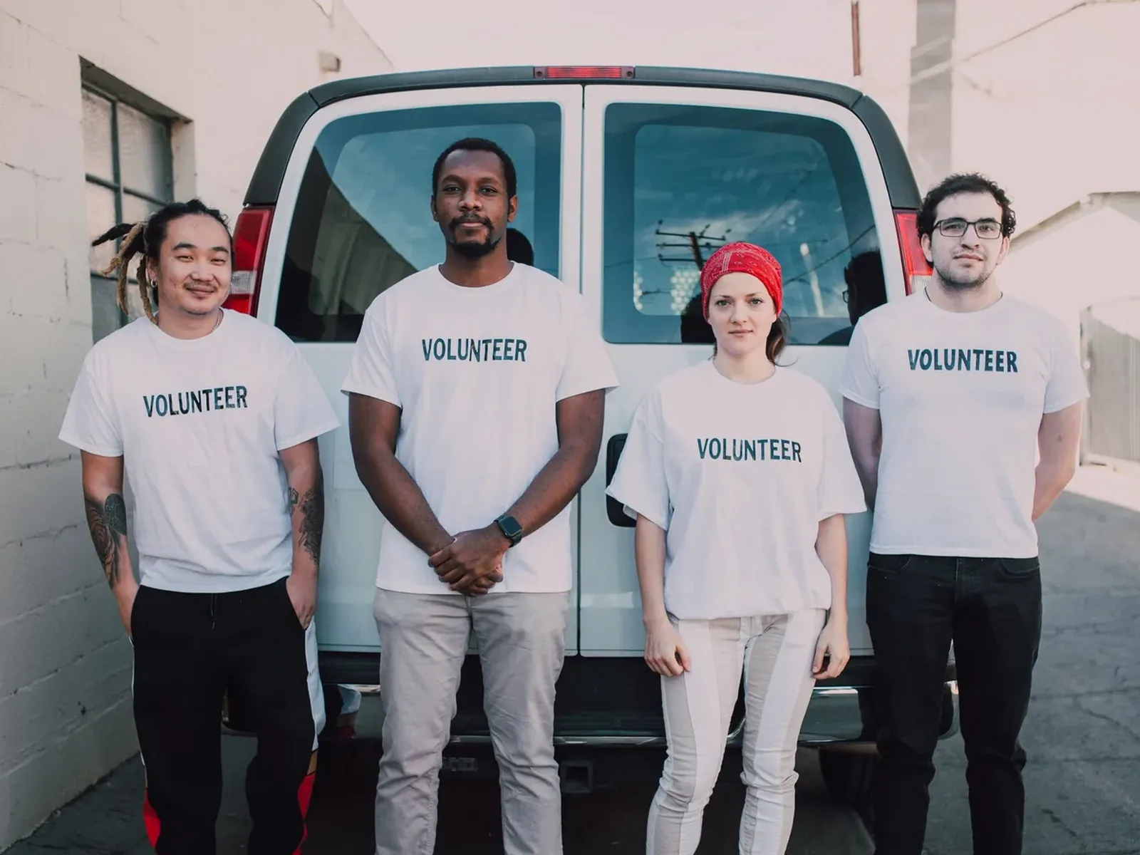 Four volunteers standing in front of a white van, all wearing matching white t-shirts printed with the word VOLUNTEER